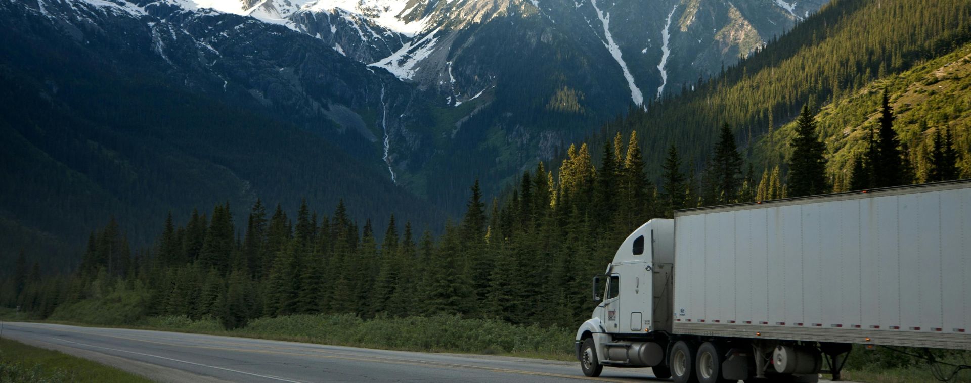 A semi-truck travels along a highway with snow-capped mountains in the background.