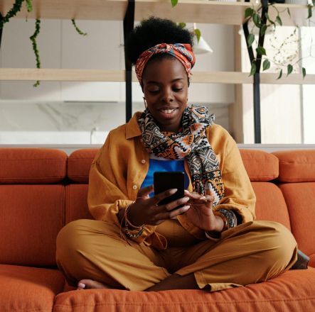 African American woman sitting on a sofa, smiling while using a smartphone in a modern indoor setting.