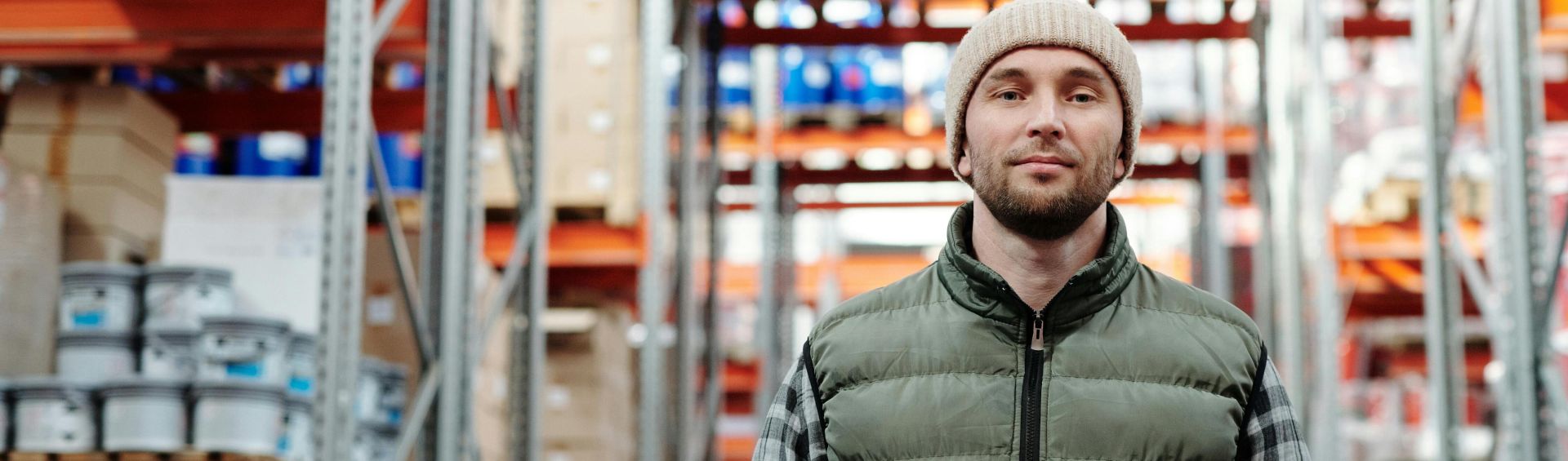 A warehouse worker wearing a beanie uses a tablet to manage inventory in a storeroom with shelves.